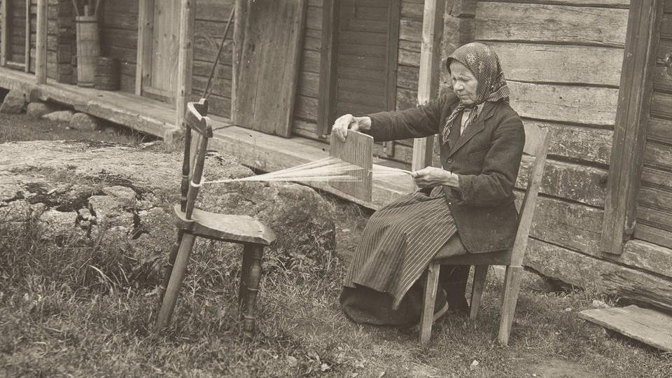 Mustavalkoinen kuva, jossa vanha nainen kutoo nauhaa käyttäen leveää nauhapirtaa.